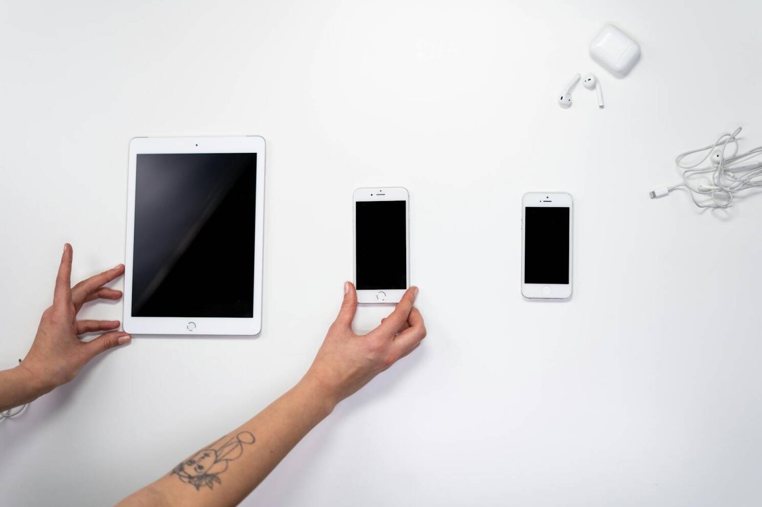 Top view of hands arranging an iPad, iPhones, and earphones on a white surface. Perfect for tech-related imagery. refurbished iPhones, quality refurbished iPhones, second-hand iPhones, iPhones for sale